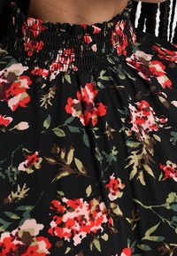 Blouse noire à col montant avec un col smocké présentant des motifs floraux rouges, roses et verts sur un tissu léger.