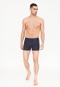 Boxers ajustados de hombre en azul marino con una cinturilla suave, diseño entallado, tela lisa y longitud hasta medio muslo. Pies descalzos hasta los tobillos, sin ropa adicional.