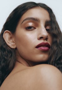 Gros plan d'une personne avec des cheveux ondulés châtain foncé, portant un rouge à lèvres baies profond, une peau mise en valeur et de petites boucles d'oreilles en or.