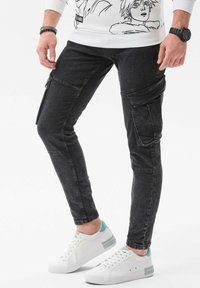 Pantalones cargo de mezclilla negra con ajuste tapered, que presentan bolsillos laterales y detalles de costura en las rodillas. Llevados con zapatillas blancas con acentos en turquesa.