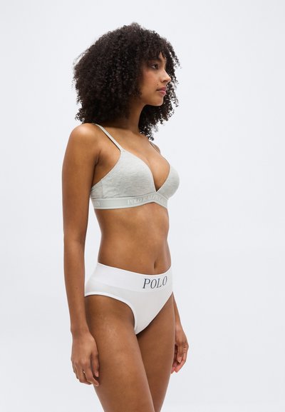 Femme aux cheveux bouclés portant un bralette Polo Ralph Lauren gris clair et une culotte taille haute blanche avec le logo "POLO" sur la ceinture, debout de profil.