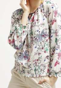 Blommig blus i lätt tyg, med vit bas och färgglada blommönster, elastisk smockad nederkant och rynkade ärmar.