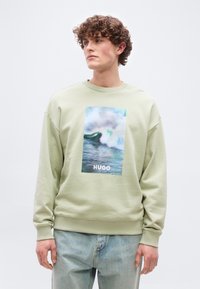 Νέος άνδρας με σγουρά μαλλιά που φοράει ανοιχτοπράσινο φούτερ με φωτογραφία κύματος και κείμενο «HUGO», σε συνδυασμό με ανοιχτό μπλε τζιν.