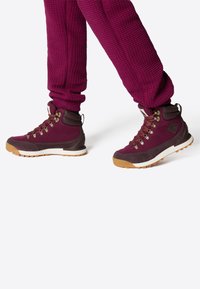 Stivali da trekking bordeaux con tessuto testurizzato, occhielli in metallo e suola in gomma marrone. Completati da pantaloni bordeaux a fantasia.