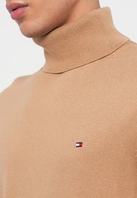 Beige korkeakauluksinen neulepaita, joka on valmistettu pehmeästä, ribatusta kankaasta. Paidassa on pieni punaisella, valkoisella ja sinisellä brodeerattu logo rinnassa.