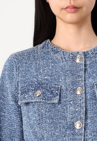 Blauwe denimjack met een gestructureerde afwerking, voorzien van zilveren knopen, twee borstzakken en een ronde halslijn.