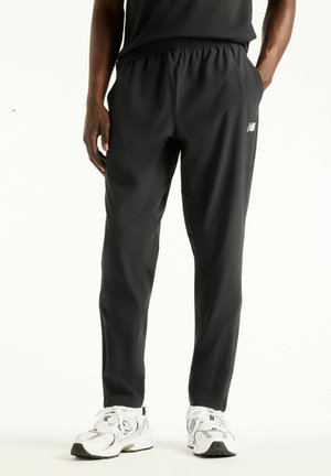 Man met zwarte New Balance sportbroek en witte New Balance sneakers staat met handen in de zakken.