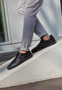 Personne portant un pantalon gris clair et des baskets noires à tige basse marchant sur une rampe en béton à l'extérieur près d'un bâtiment.