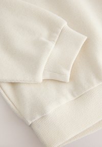 Dettaglio di un maglione color crema con tessuto testurizzato, polsini e orlo a coste. Superficie liscia con un motivo sottile.