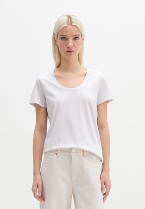 Młoda kobieta o prostych blond włosach, ubrana w prostą białą koszulkę i jasnobeżowe spodnie, stoi na białym tle.