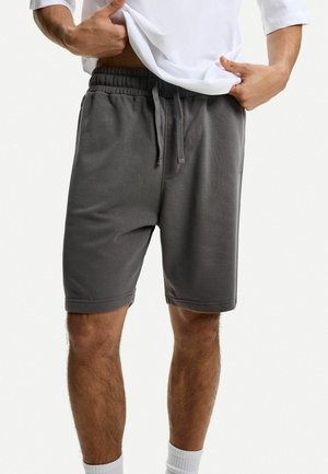 Mann trägt dunkelgraue Sweatshorts knapp über dem Knie mit elastischem Bund und Kordelzug, hebt leicht ein weißes Hemd an, untere Beine sichtbar.