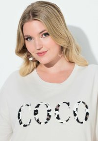 Kremfarvet T-shirt med "COCO" i sort og brun dyreprint. Løs pasform, rund halsudskæring, blødt bomuldsstof og korte ærmer.