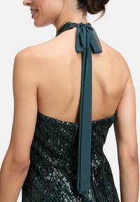 Frau in einem dunkelgrünen, mit Pailletten besetzten Halterkleid, das am Hals mit einer Schleife gebunden ist, und ihren nackten Rücken sowie das Seitenprofil ihres Gesichts zeigt.