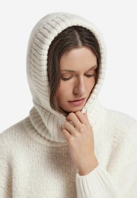 Biały dziany sweter z kapturem, o fakturowanej powierzchni i miękkim, grubym kołnierzu. Palec delikatnie dotyka tkaniny.