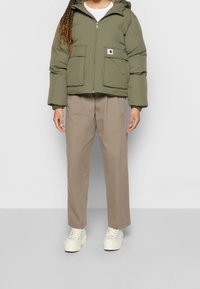 Olivengroene pufferjas met een rits, twee voorzakken en een klein logopatch; gecombineerd met beige wijde broeken en witte sneakers.