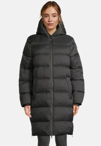 Femme portant un long manteau rembourré noir avec capuche, fermeture éclair à l'avant et poches latérales, debout sur un fond uni.