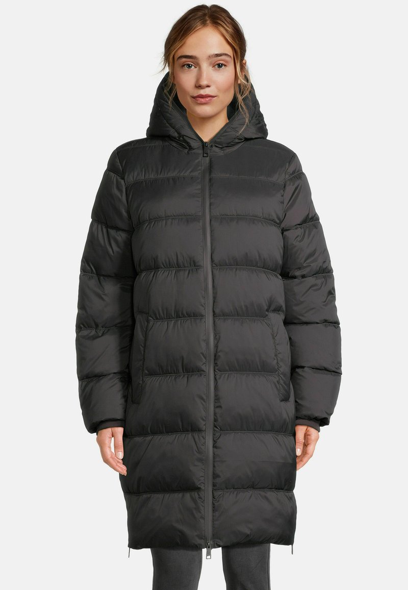 Femme portant un long manteau rembourré noir avec capuche, fermeture éclair à l'avant et poches latérales, debout sur un fond uni.