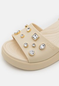 Sandale slide Crocs beige avec une large sangle, décorée de strass transparents et d'accents dorés. Semelle intérieure texturée et semelle durable.