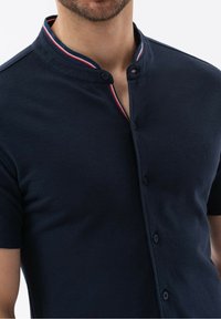 Polo de manga corta azul marino con cuello alzado, tapeta de dos botones y acentos a rayas rojas y blancas en el interior del cuello. Tejido elástico.