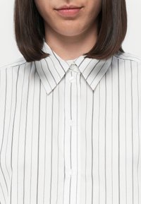 Camicia bianca con sottili righe nere verticali, con chiusura a bottone frontale e una superficie liscia e lucida.