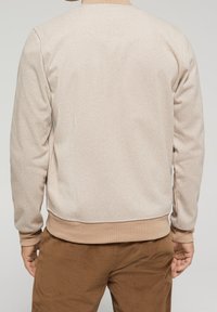 Beige herringbone-patterned sweatshirt with ribbed cuffs and hem, made from textured fabric, worn with brown corduroy pants.