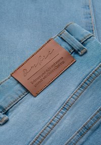 Jean en denim bleu clair avec une étiquette en cuir marron cousue sur la ceinture portant l'inscription "Ombre Clothing" et un slogan de la marque.