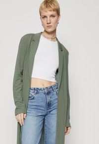 Cardigan lung, deschis în față, de culoare verde măsliniu, făcut dintr-un material moale, purtat peste un top alb scurt și blugi albaștri cu talie înaltă, având o croială lejeră.
