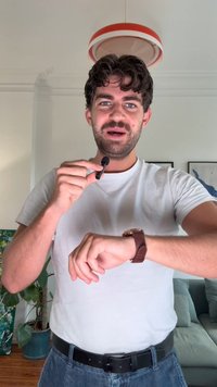 Čierny klipsňový mikrofón držený v pravej ruke, oblečený s bielym tričkom, so hnedým koženým náramkovým hodinami na ľavom zápästí.