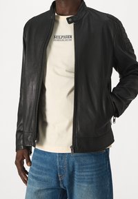 Hombre con chaqueta de cuero negra sin abrochar, camiseta gráfica crema de Hilfiger y jeans azules, de pie con la mano en el bolsillo.