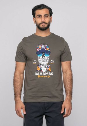 Homme portant un t-shirt vert olive avec un graphique coloré de crâne en lunettes de soleil et casquette, texte "Bahamas Beach for life", et un pantalon foncé.