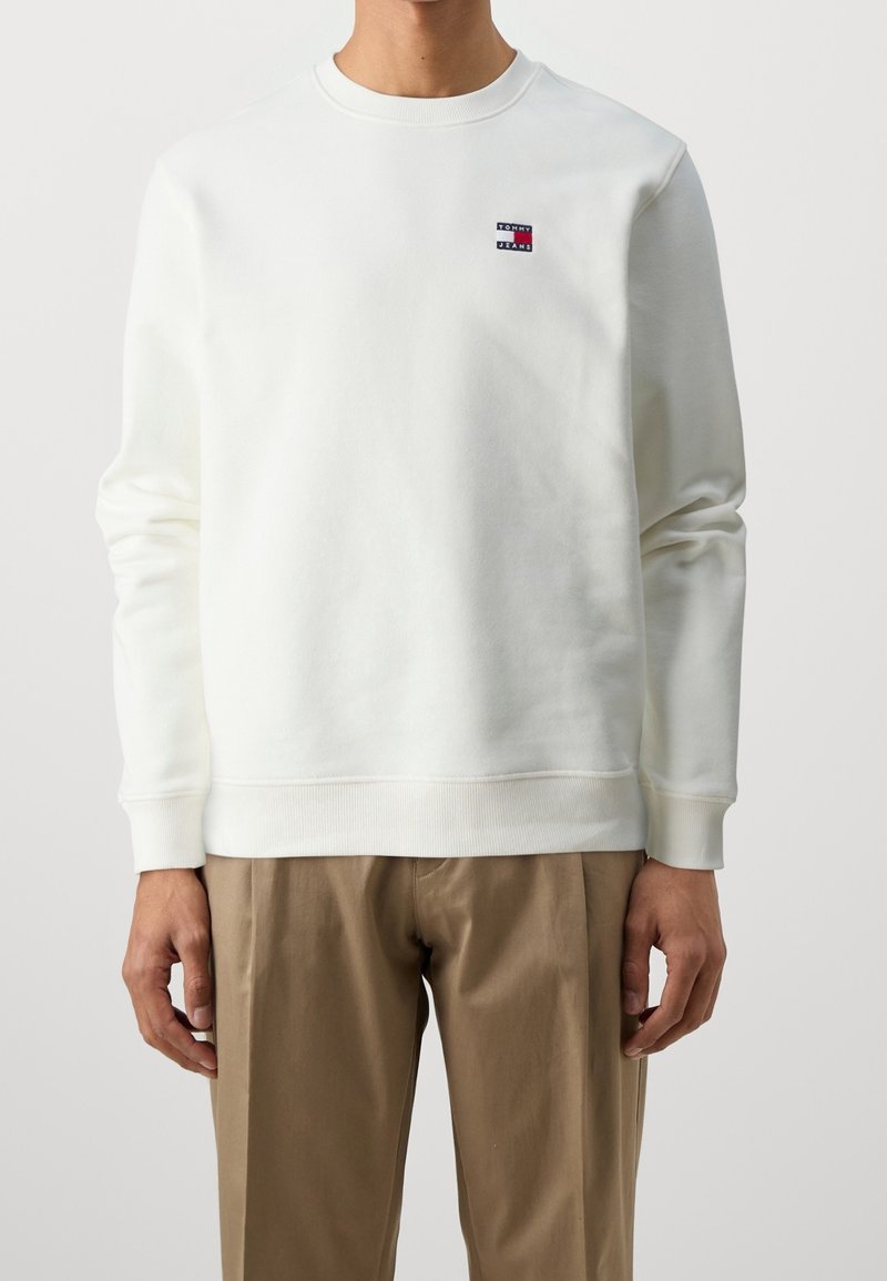 Bílý sweatshirt s kulatým výstřihem z bavlny. Na levé hrudi má malou nášivku s logem v červené a modré barvě a žebrované manžety a lem.