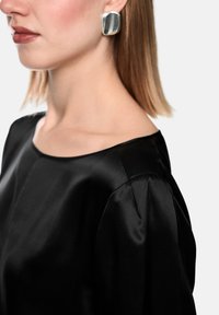 Woman wearing a black satin blouse and large silver square earrings, with straight shoulder-length hair against a white background.