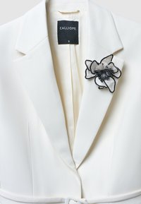 Weißer Blazer mit strukturiertem Design, versehen mit einem schmalen Kragen und einer abnehmbaren schwarz-grauen Blumenbrosche. Innenseite in hellem Creme.