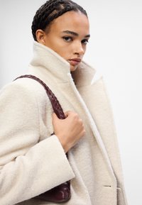 Bílý vlněný kabát s vysokým límcem a texturovanou tkaninou, doplněný burgundskou koženou taškou se zapleteným popruhem.