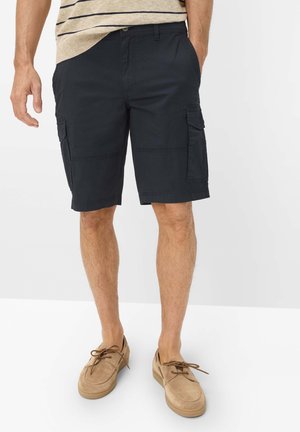 Man draagt marineblauwe cargoshorts met zijzakken, een beige gestreept overhemd en beige casual veterschoenen, staand met handen bij de zakken.