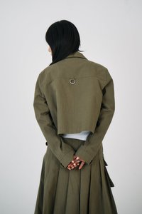 Person med langt svart hår, iført en olivengrønn jakke og plisséskjørt, står med hendene foldet bak ryggen mot en ensfarget bakgrunn.