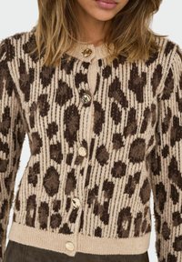 Žena u bež kardiganu s pahuljastim tamnosmešim leopard printom i sjajnim bež dugmadi, prikazana od grudnog koša do bokova.