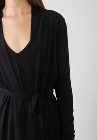 Černá kimono stylová župan vyrobená z měkké, lehké tkaniny. Má opasek v pase, dlouhé rukávy a hluboký výstřih do V.