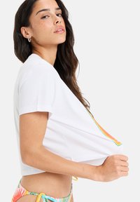 White cropped t-shirt with short sleeves and a colorful graphic design on the front, worn over a multi-colored bikini.