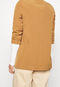 Hnědý přizpůsobený blazer s hladkou texturou, propracovanými rameny a ohrnutými manžetami, doplněný o bílou košili s dlouhým rukávem pod ním.