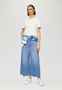 Polo shirt blanc à manches courtes, associé à un jean en denim bleu clair à jambes larges. Un pull bleu et blanc à motif noué à la taille. Baskets blanches.