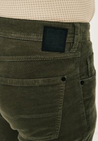 Olivgrüne Hose mit strukturierter Oberfläche, ausgestattet mit einem schwarzen Etikett am Bund und mehreren Taschen mit kontrastierenden Nähten.