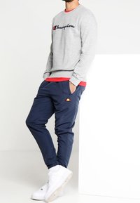 Grauer Hoodie mit "Champion"-Logo, über einem roten Shirt getragen. Marineblaue Jogginghosen mit Logodetails, kombiniert mit weißen Sneakers. Einfaches und lässiges Design.