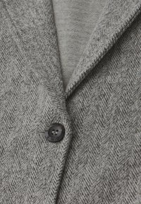 Grå herringbone-blazer med en texturerad yta och svarta knappar som detalj. Kragen har en notched-design som visar upp lager av material.