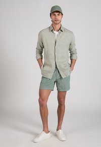 Hombre con gorra verde, camisa gris claro abotonada, pantalones cortos verdes y zapatillas blancas, de pie con las manos en los bolsillos.