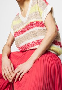 Neulotut lyhythihaiset neuleet, joissa on keltaisia, punaisia ja vaaleita vaakaraitoja vaalealla taustalla; yhdistetty vaaleanpunaiseen laskostettuun hameeseen.