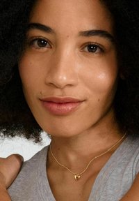 Collier en pendentif cœur en or sur une chaîne délicate, porté sur un haut en tissu gris, avec des teintes de peau douces et naturelles et des cheveux noirs bouclés encadrant le visage.