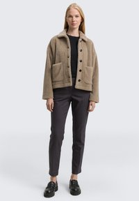 Beige fleece jacket with a collar, two front pockets, and black buttons, paired with dark trousers and black loafers. Smooth texture.