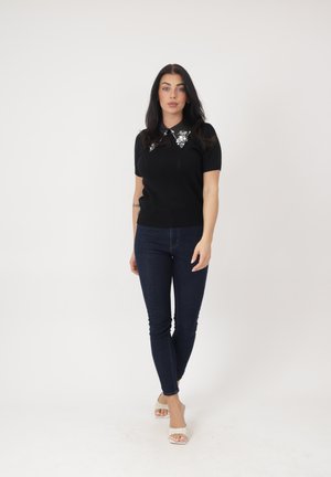 Frau steht und trägt ein schwarzes kurzärmeliges Oberteil mit verziertem Kragen, dunkle Skinny-Jeans und weiße Sandaletten mit Absatz vor einem schlichten weißen Hintergrund.