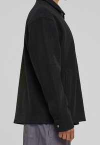 Svart jacka med lång ärm, krage, sidfickor och avslappnad passform. Tillverkad av ett mjukt material med en slät textur.